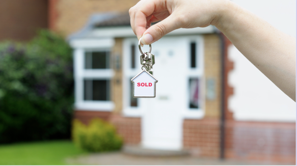 Image of a hand holding a key with a charm that says “sold” in front of a brand new home.