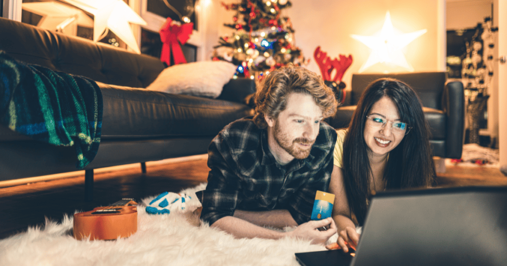 Couple is lying on the floor during Christmas time with a Christmas tree behind them, shopping for a home on a laptop.