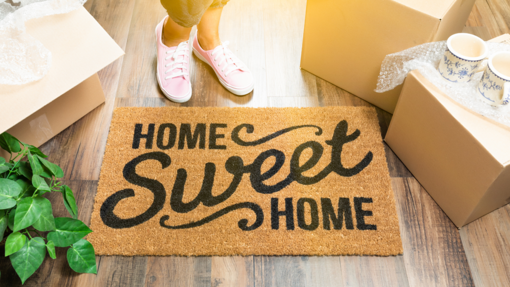 Welcome mat that says “Home Sweet Home” placed in the doorway of a new home with a person standing nearby surrounded by moving boxes.
