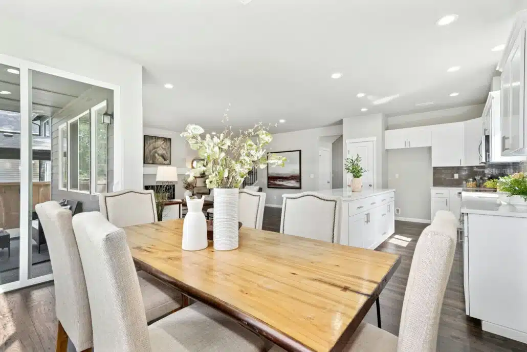 A wood kitchen table with tan chairs around it. In the background there is a kitchen and living room visible.