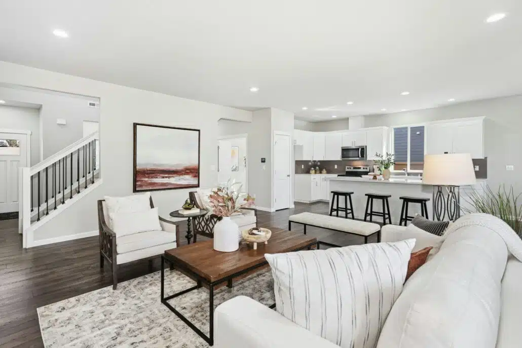 A modern living room with wood floors and white furniture. In the background a kitchen and island are visible.