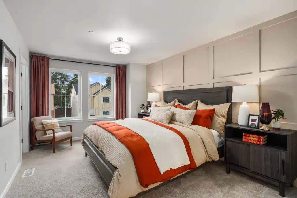 A carpeted bedroom with a tan accent wall. There are orange decorative pillows on the bed and a chair in the corner.