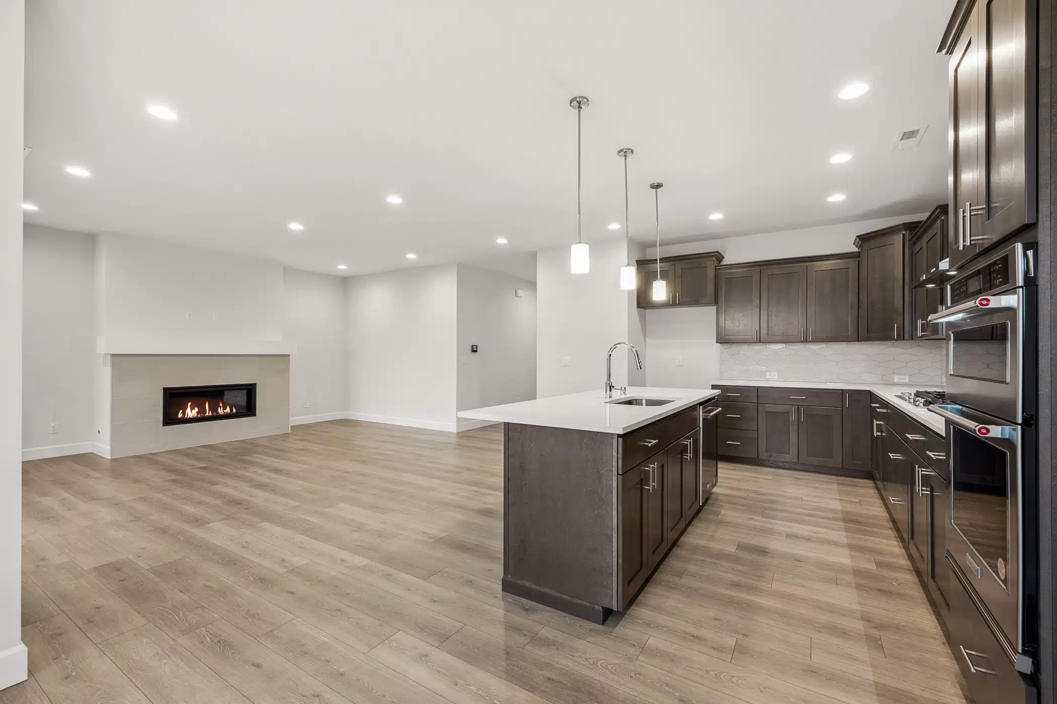 A modern kitchen with dark wood cabinets and an island. An empty living room with a fireplace is behind it.