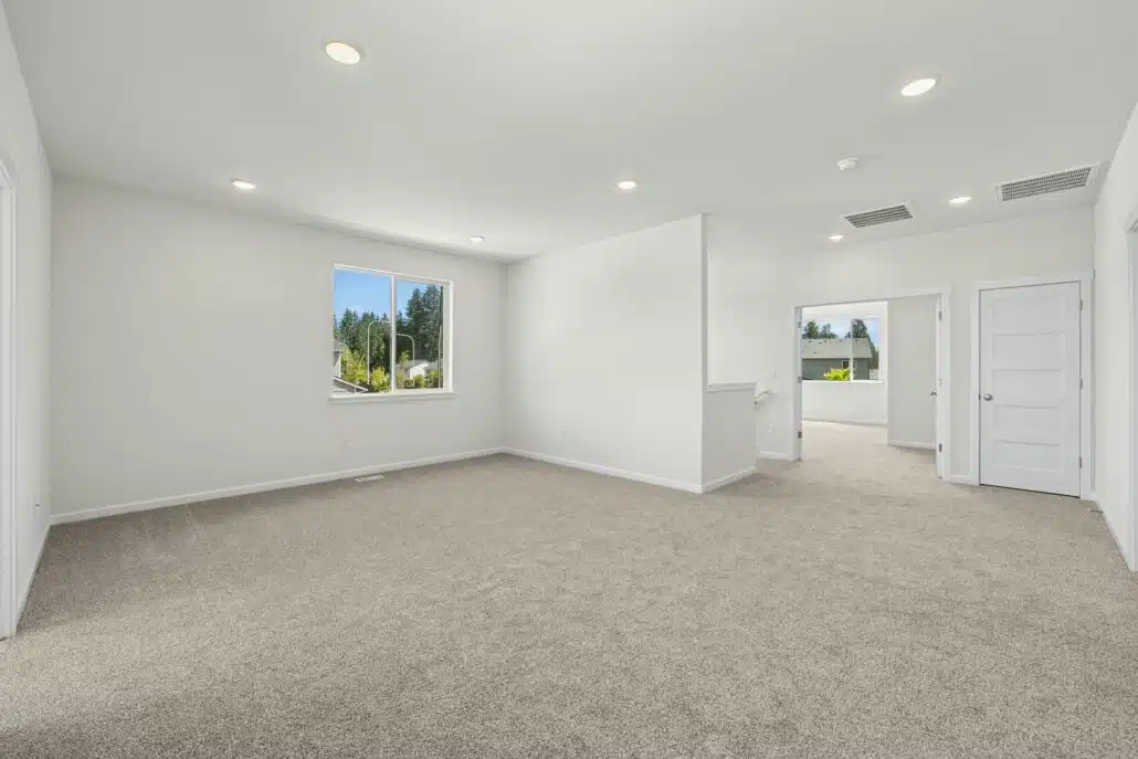 A large, empty, carpeted room with white walls. There is a small window in the corner.