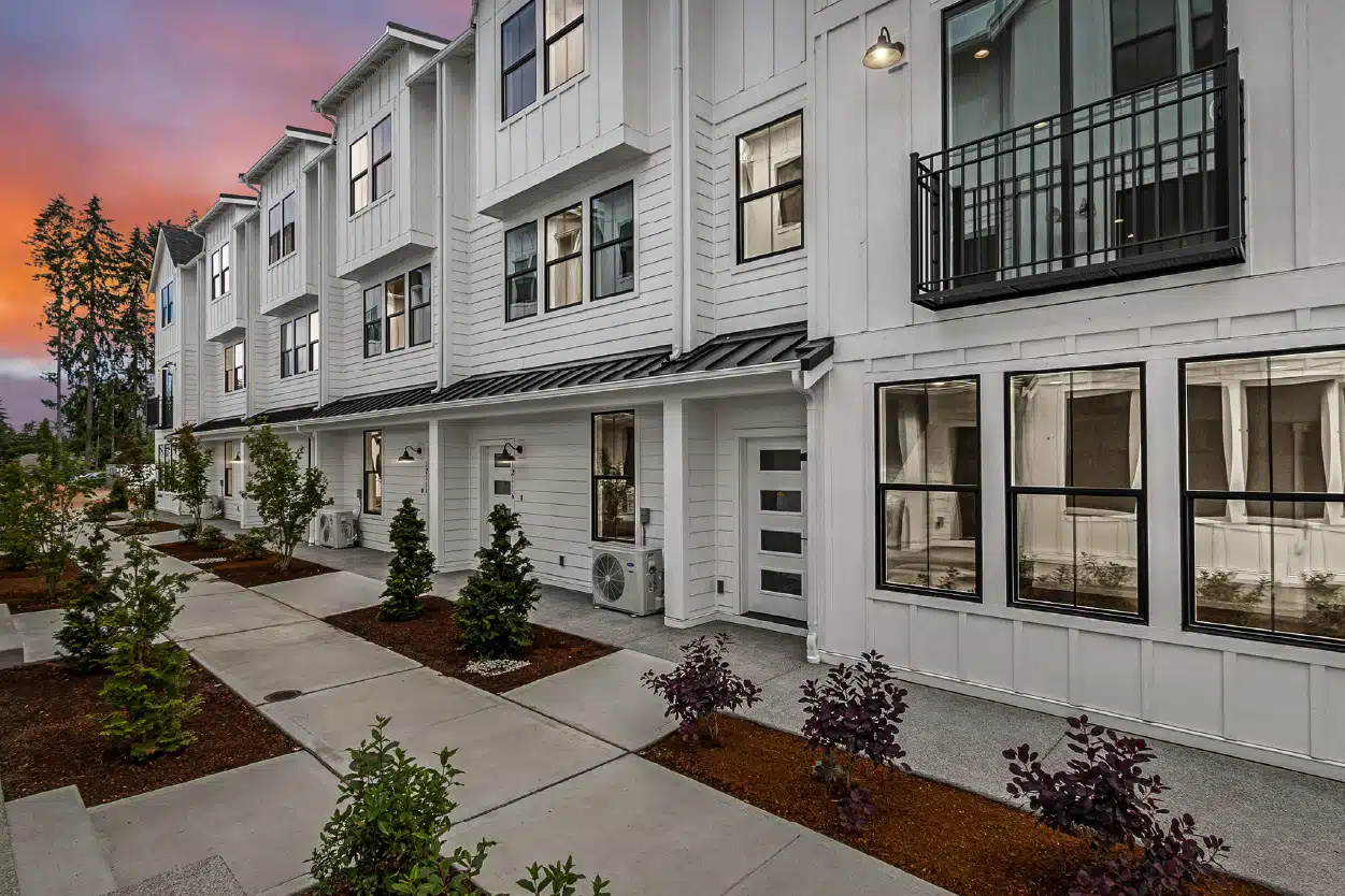 A row of modern white townhomes with black window trim and metal awnings, featuring small landscaped front yards and a smooth concrete walkway under a vibrant sunset sky.