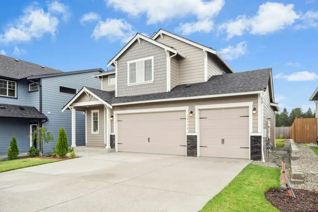 Two-story house with gray siding, a three-car garage, and a concrete driveway under a bright blue sky.