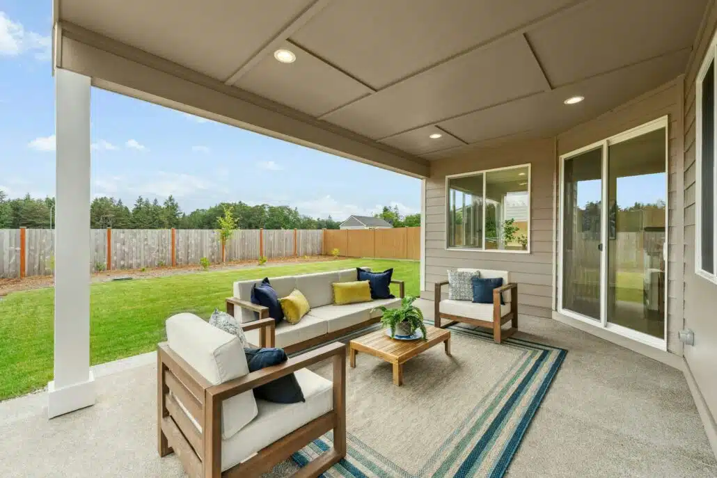 Covered backyard patio with outdoor seating including a sofa, two armchairs, and a coffee table on a rug. The space overlooks a grassy yard enclosed by a wooden fence, with trees and houses in the background under a blue sky.