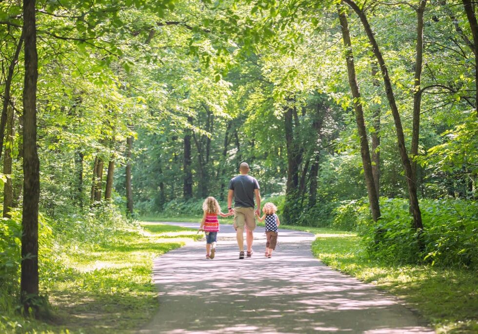 Rear view of a father and his two young daughters going for a walk on a paved trail through the wooded park. The three are walking in a row holding hands with each other. Sunlight is coming through the open spaces, so the trail is partially shaded and partially in sunlight.